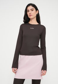 Καφέ μακρυμάνικη μπλούζα με ραβδώσεις, με στρογγυλή λαιμόκοψη και διακριτικό λογότυπο της μάρκας σε ανοιχτό ροζ, συνδυασμένη με φούστα σε ανοιχτό ροζ.