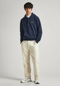 Sudadera navy con bolsillo canguro y cordón, combinada con pantalones beige claros y zapatillas grises con acentos negros y superficies texturizadas.