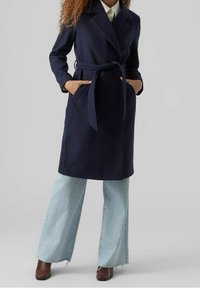 Cappotto belted in misto lana blu navy con ampie revers, tasche frontali e vestibilità sartoriale, abbinato a jeans a gamba larga in denim chiaro e scarpe marroni.