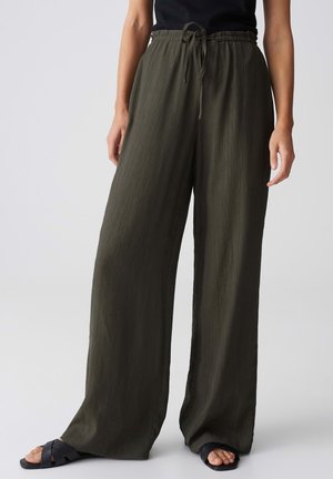 Femme portant un pantalon ample vert olive à jambes larges avec une taille à cordon et des sandales noires, debout devant un fond clair uni.