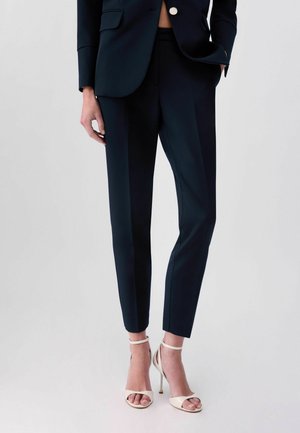 Donna che indossa pantaloni neri su misura alla caviglia e un blazer abbinato, abbinati a sandali bianchi con tacco alto e cinturini alla caviglia.