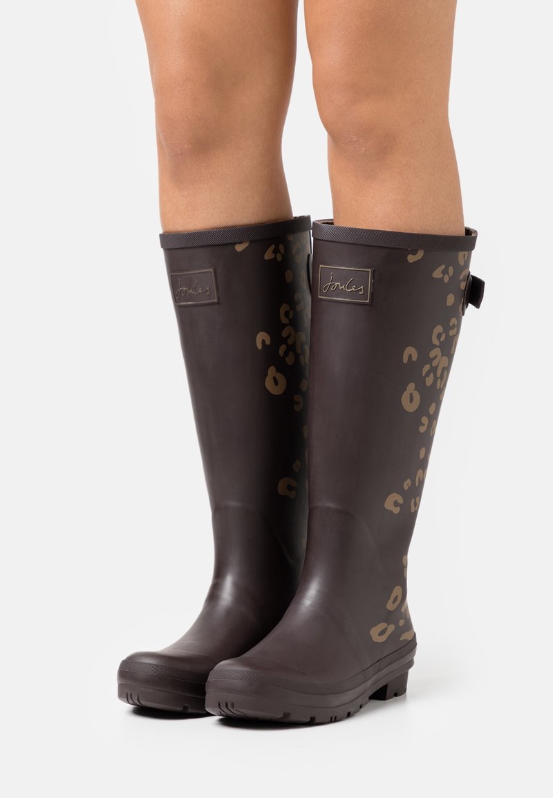 Bottes de pluie en caoutchouc brun foncé montantes aux genoux avec des taches léopard brun clair, portées sur des jambes nues sur fond blanc.