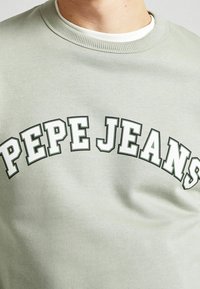 Ljusgrön sweatshirt med ribbad rund halsringning, som har kraftig vit och mörkgrön text "PEPE JEANS" i en bågformad design över bröstet.