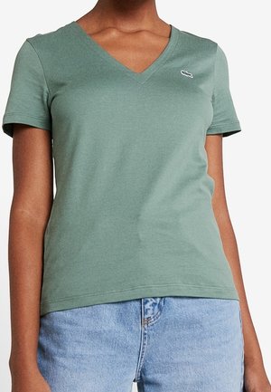 Kobieta ubrana w zielony T-shirt z krótkim rękawem w serek z małym białym logo na piersi, zestawiony z jasnoniebieskimi jeansami.