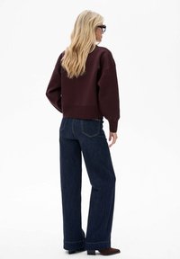 Bordowy krótki sweter z opuszczonymi ramionami, zestawiony z szerokimi ciemnymi dżinsami o prostych nogawkach. Gładka faktura i prosty fason, z widocznymi przeszyciami.