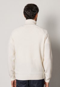 Vit stickad tröja med hög krage, ribbade muddar och linning. Mjuk textur och rent design, bärs över mörka jeans.