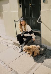 Jeune femme en casquette noire et pull assise sur des marches avec une tasse de café, tenant une laisse à côté d'un petit chien brun duveteux sur le trottoir.