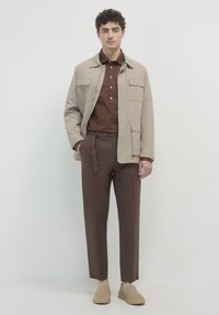 Junger Mann in einer beigefarbenen Jacke über einem braunen Hemd mit Knöpfen, braunen Hosen mit einem Gürtel und beigefarbenen Slippern vor einem schlichten Hintergrund.