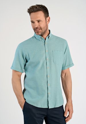 Man in een turkooizen overhemd met korte mouwen en borstzak, en een donkere broek, staand met één hand in de zak.