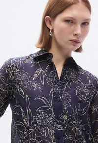 Camisa floral azul-marinho com design de gola, apresentando padrões florais em creme, fecho de botões e textura leve e transparente.