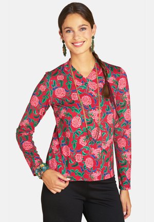 Woman with brown braided hair wears bright red floral long-sleeve top, black pants, long necklace, green earrings, and a beaded bracelet.