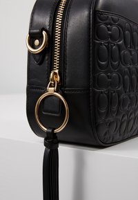 Musta nahkalaukku, jossa on kuvioitu, kohokuvioitu design, kultainen metalliosat, vetoketjusuljenta ja tupsukoriste, joka on kiinnitetty kultarenkaaseen.