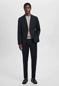 Traje azul marino a medida con un blazer estructurado y pantalones de corte slim, combinado con un suéter beige y mocasines negros. Tejido suave, acentos mínimos.