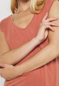 T-shirt rosa pallido a V con maniche corte, realizzato in tessuto morbido. Presenta una vestibilità comoda con una superficie liscia. Braccia incrociate davanti.