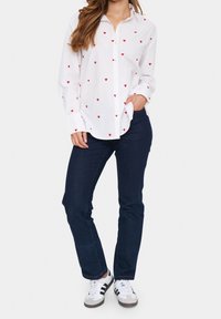 White button-up shirt with red heart patterns, paired with dark blue jeans and white sneakers featuring black stripes. Casual fit.