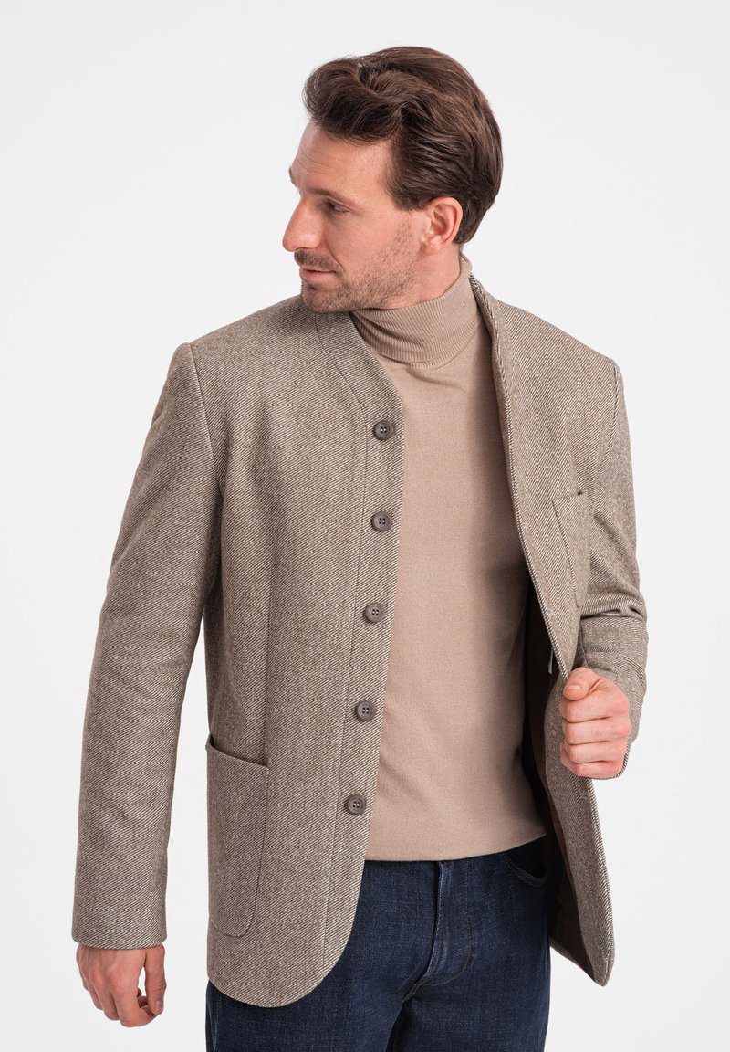 Brauner Fischgrätenblazer mit hohem Kragen, sechs Knöpfen und zwei Seitentaschen, kombiniert mit einem beigen Rollkragenpullover und dunkelblauen Jeans.