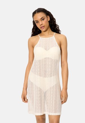 Mujer con cabello rizado que lleva un vestido blanco sin mangas de estilo crochet con tirantes finos, posando frente a un fondo claro y liso.