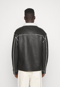 Uomo di spalle che indossa una giacca di pelle nera con colletto di shearling bianco e cuciture bianche, abbinata a pantaloni off-white.