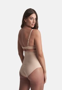 Donna che indossa lingerie modellante beige a vita alta e reggiseno coordinato, che guarda oltre la spalla contro uno sfondo chiaro e semplice.