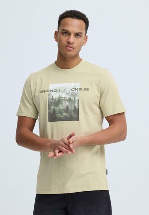 Ung mand med kort krøllet hår iført en beige t-shirt med et skovmotiv og teksten "BLEND", stående foran en ensfarvet lys baggrund.