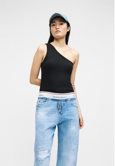 Νεαρή γυναίκα που φοράει μαύρο τοπ με έναν ώμο, ανοιχτό μπλε τζιν με σκισίματα και λευκή ελαστική μέση, καθώς και ανοιχτό μπλε καπέλο από τζιν.