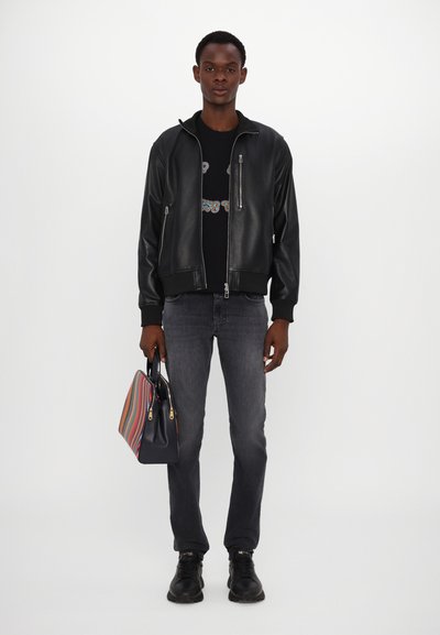 Μαύρη δερμάτινη τζάκετ βομβιστή, μαύρο μπλουζάκι με γραφικά, γκρι τζιν, μαύρα αθλητικά παπούτσια, κρατώντας μια ριγέ τσάντα με μπλε, κόκκινες και κρεμ αποχρώσεις.