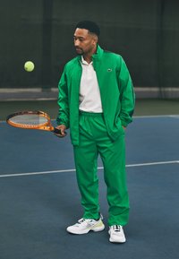 Zöld sportos szabadidőruha cipzáras dzsekivel és gumis derekú nadrággal, fehér pólóval és fehér sportcipővel kombinálva, teniszütőt tartva.