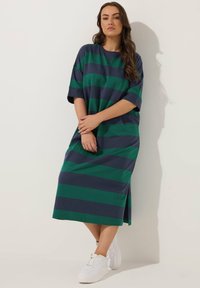 Nainen seisoo valkoista seinää vasten yllään tummansininen ja vihreä leveäraitainen midimekko sekä valkoiset lenkkarit, kädet ristissä edessä.