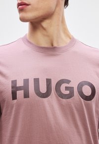 Svetloružová bavlnená tričko s veľkým čiernym nápisom "HUGO" na prednej strane. Okrúhly výstrih a krátke rukávy s mäkkou textúrou.