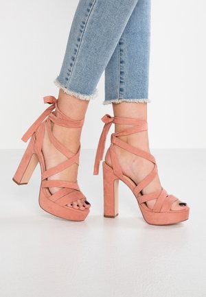 Nærbillede af fødder iført lyserøde ruskinds højhælede sandaler med ankelremme, der er bundet i sløjfer, sammen med cropped lyseblå jeans med frynset kant.