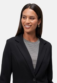 Blazer nero con spalle strutturate e chiusura a un bottone, indossato sopra un top con motivo testo in bianco e nero.