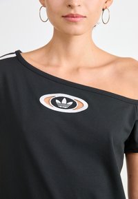 Juoda off-shoulder marškinėliai su balta siuvinėta logo ovalo formos išpjovoje. Minkšta medžiaga, laisvas kirpimas ir trumpi rankoviai.