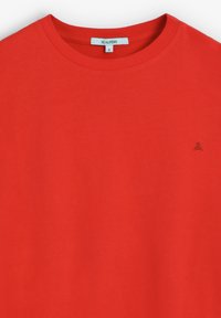 Camiseta de algodón roja con escote redondo y mangas cortas. Presenta un pequeño logotipo sutil en el pecho. Textura suave, diseño básico.