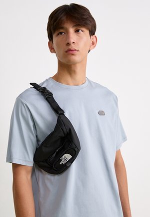 Jeune homme portant un t-shirt bleu clair et un sac bandoulière noir The North Face, regardant légèrement vers le haut sur un fond uni.