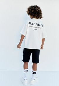 Bílý tričko s krátkým rukávem s černým nápisem "ALLSAINTS" na zádech, v kombinaci s černými kraťasy a bílými teniskami s černými pruhy na ponožkách.