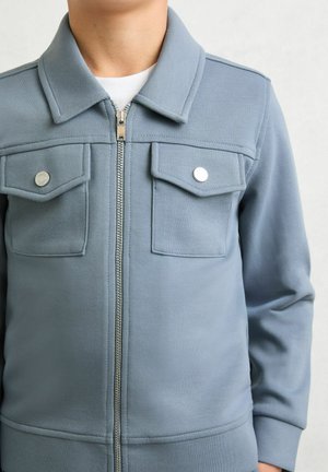 Jasnoniebieska kurtka na zamek z kołnierzykiem, dwoma kieszeniami na piersi i srebrnymi zatrzaskami. Materiał jest gładki, o casualowym kroju.