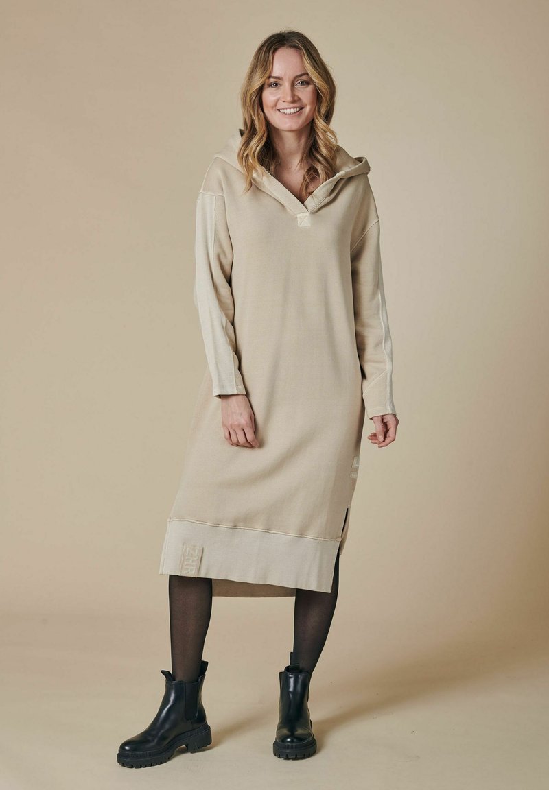 Beiges Hoodie-Kleid mit langen Ärmeln, strukturierten Paneeldetails, Seitenschlitzen und bestickten Akzenten, kombiniert mit schwarzen Knöchelstiefeln.