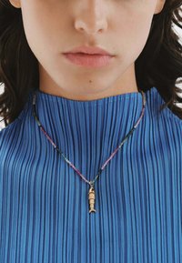 Collar con pendiente de pez dorado y cadena de cuentas multicolores sobre una blusa azul con textura y plisados, que presenta finos pliegues y una tela suave.