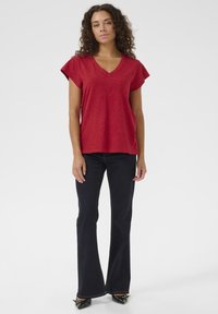 Červený top s výstřihem do V a krátkým rukávem z texturované látky, spárovaný s tmavě modrými zvonovými džíny a černými botami na podpatku. Jednoduchý, neformální styl.