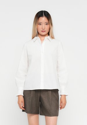 Vrouw in een witte overhemd met knopen en gerimpelde manchetten, met een bruine op maat gemaakte broek tot de knie tegen een plain witte achtergrond.
