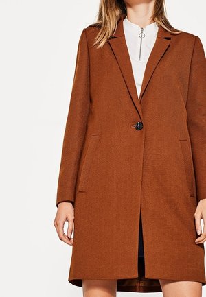 Nainen pukeutuneena keskipitkään ruosteenruskeaan yksiriviseen takkiin, jossa on yksi nappi, valkoisen topin päällä, jossa on hopeinen vetoketjun vetorenkaan kauluksessa.