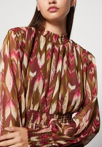 Mönstrad blus i flera färger med hög krage, långa ärmar och resårmidja. Tyget har ett blomster-i-kat-mönster i rosa, brunt och gräddvitt.