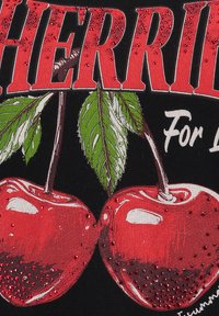 Una tela negra presenta un gráfico de dos brillantes cerezas rojas con hojas verdes. "CEREZAS" está impreso de manera prominente en rojo negrita arriba, con ornamentaciones.