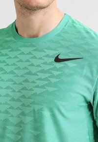 Camiseta deportiva verde con un patrón triangular texturizado, cuello redondo y un logotipo negro de Nike en la parte superior izquierda.