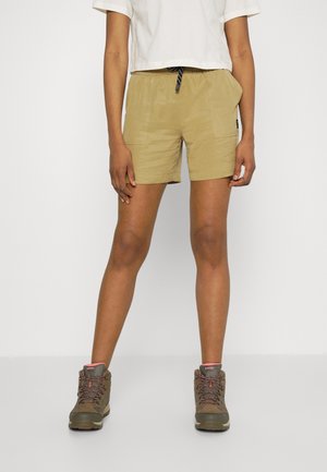 Persona que lleva pantalones cortos beige con bolsillos delanteros, un top corto blanco y botas de senderismo marrones, de pie frente a un fondo liso.