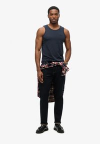 Top sin mangas azul marino, jeans oscuros, camisa de cuadros atada a la cintura y mocasines negros. Material de algodón con una silueta ajustada y costuras visibles.