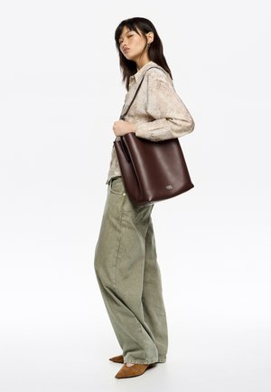 Bolsa tote de cuero marrón con una textura suave, que presenta una base plana y una sola correa para el hombro. Modelo vistiendo pantalones verdes holgados.