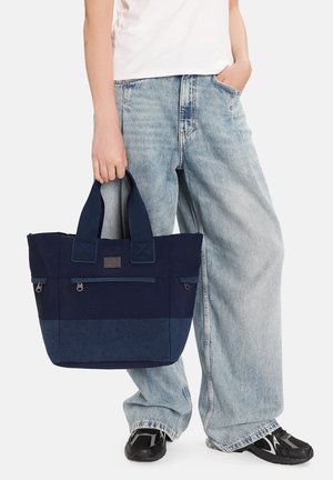 Persona con vaqueros anchos de pierna color azul claro y camisa blanca sosteniendo un bolso tote azul marino con bolsillos con cremallera por las asas.