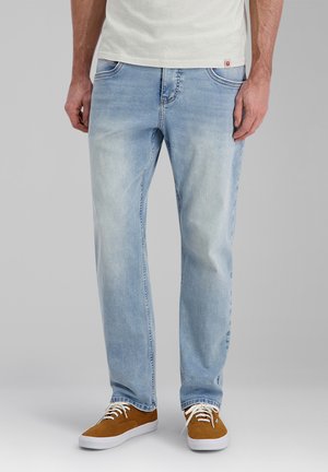 Mann mit hellblauen Jeans und braunen Schnürsneakern steht vor einem schlichten Hintergrund.