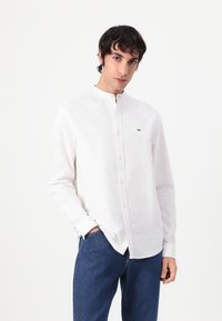 Weißes Hemd mit Stehkragen, aus glattem Baumwollstoff gefertigt, mit kleinem Logo auf der Brust. Getragen mit blauen Jeans.